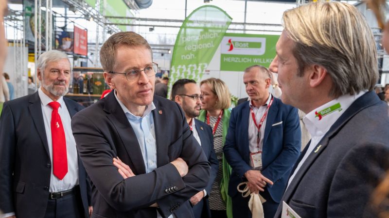 Mehrere Personen in Businesskleidung stehen in einer Messehalle vor einem grünen Banner mit der Aufschrift 'Herzlich Willkommen' und führen Gespräche.