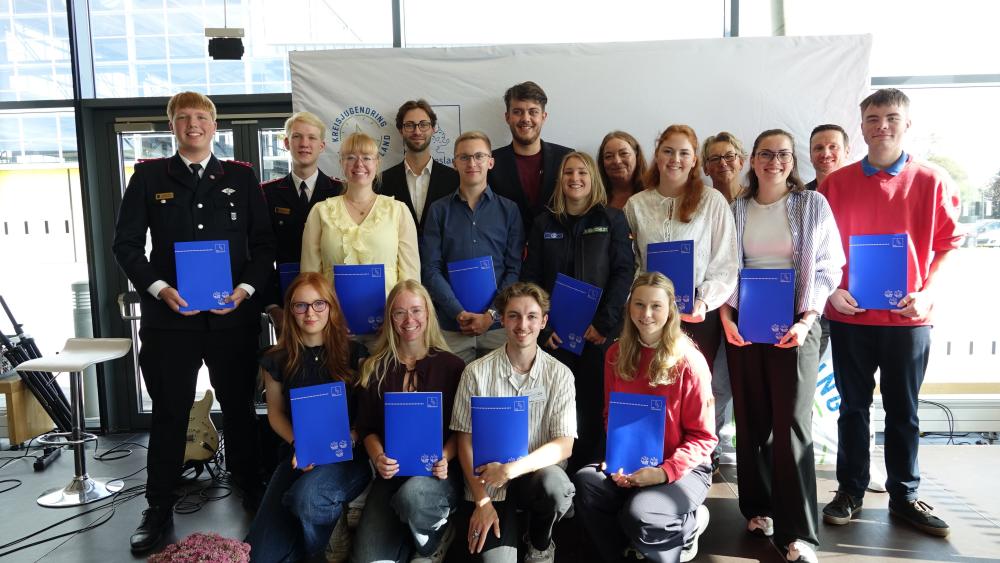 Gruppe von Menschen unterschiedlichen Alters steht in zwei Reihen vor einem hellen Hintergrund und hält blaue Ordner in den Händen