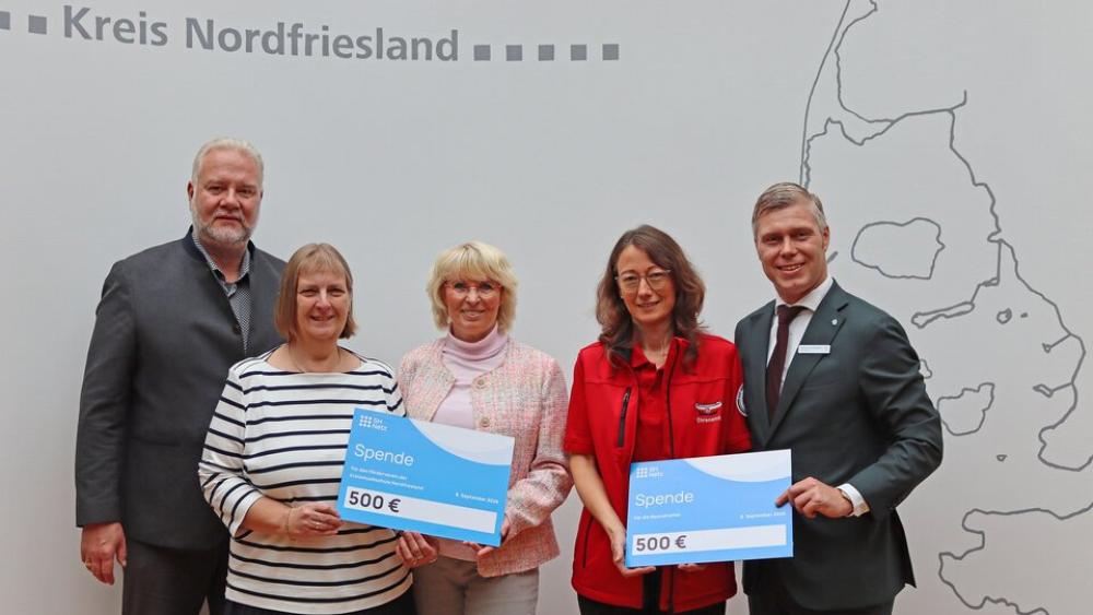 Fünf Personen stehen vor einer Wand mit der Aufschrift 'Kreis Nordfriesland' und einer Landkarte, zwei Frauen halten jeweils einen Scheck über 500 Euro