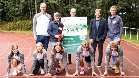 Fünf Jugendliche in Startposition auf einer Laufbahn vor vier stehenden Erwachsenen, die ein Plakat mit der Aufschrift 'Achtung, fertig, los!' halten, im Hintergrund Bäume.