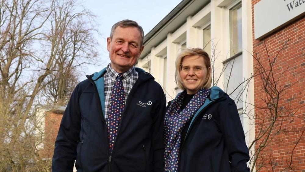 Zwei Personen in dunklen Jacken mit blauen Kapuzen und gemusterten Hemden stehen vor einem Backsteingebäude mit weißen Fensterrahmen und kahlen Bäumen im Hintergrund.