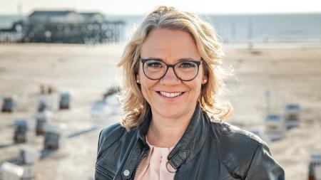 Eine blonde Frau. Im Hintergrund unscharf der Strand von St. Peter-Ording.