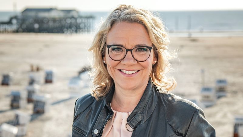 Eine blonde Frau. Im Hintergrund unscharf der Strand von St. Peter-Ording.