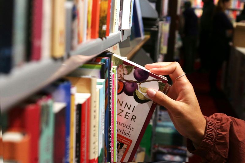 Hand mit silbernem Ring zieht ein Buch mit lila Pflaumenmotiv aus einem Bücherregal in einer Buchhandlung.