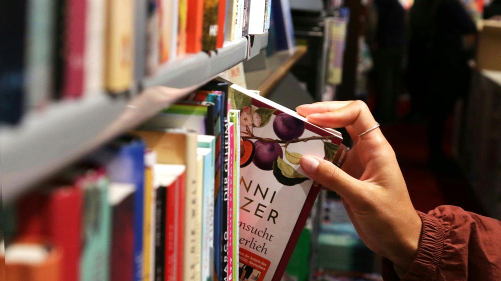 Hand mit silbernem Ring zieht ein Buch mit lila Pflaumenmotiv aus einem Bücherregal in einer Buchhandlung.