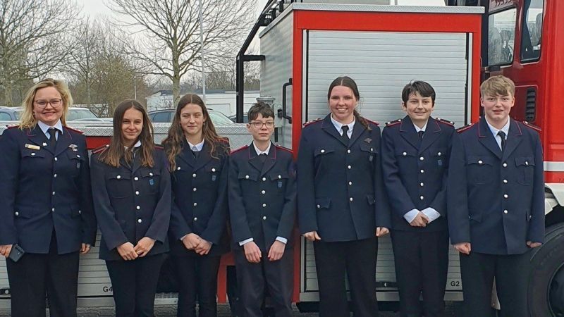 Jugendliche stehen vor einem Leiterwagen der Feuerwehr. Sie tragen blaue Feuerwehruniformen.