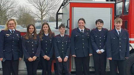 Jugendliche stehen vor einem Leiterwagen der Feuerwehr. Sie tragen blaue Feuerwehruniformen.
