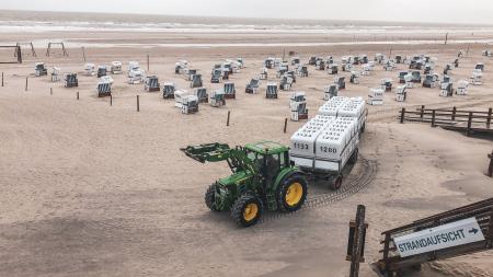 Traktor zieht Anhänger mit weißen Strandkörben auf sandigem Strand mit vielen weiteren Strandkörben im Hintergrund.