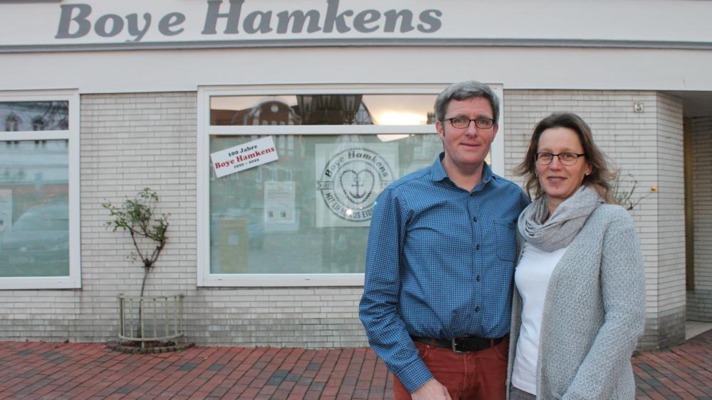 Ein Mann und eine Frau vor einer hellen gemauerten Fassade. Auf dem Gebäude im Hintergrund steht Boye Hamkens