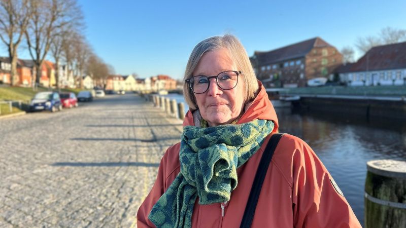 Person mit rotem Mantel und grün gemustertem Schal steht an einem gepflasterten Ufer mit Wasser und Gebäuden im Hintergrund