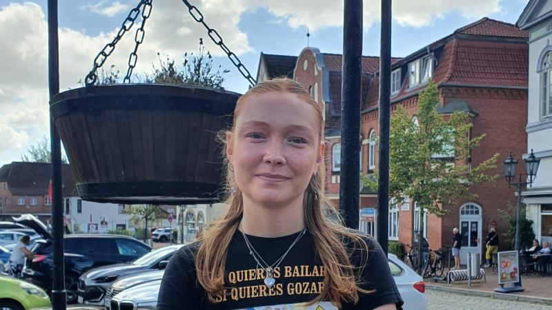 Junge Frau mit verschränkten Armen vor einem Steinbrunnen in einer belebten Altstadt mit Backsteingebäuden und geparkten Autos in Tönning.