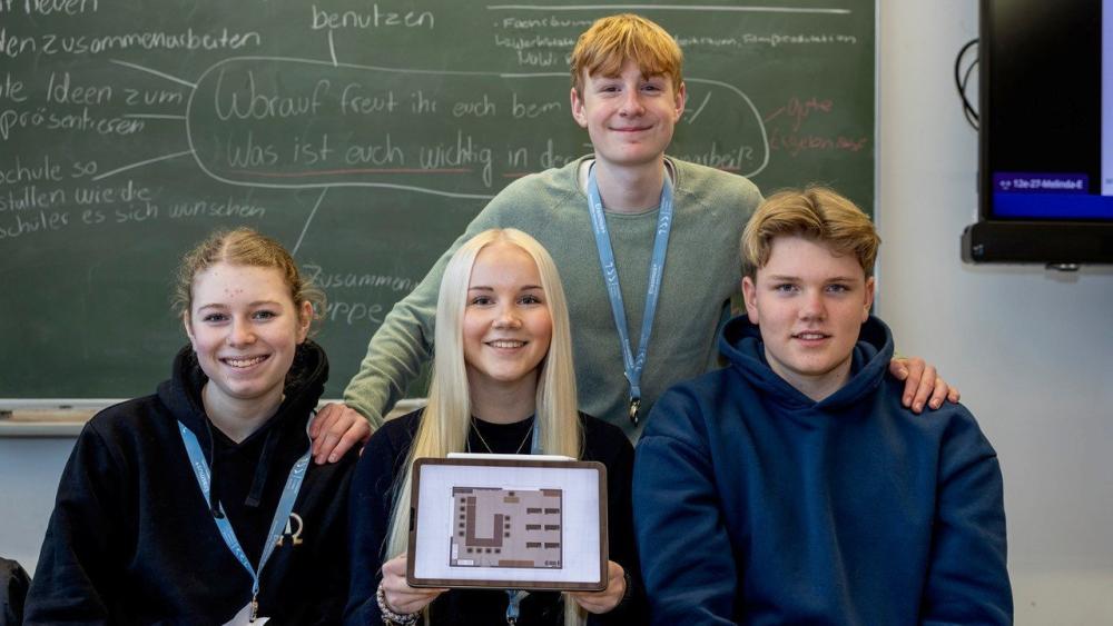 Vier Jugendliche sitzen und stehen vor einer Tafel mit handgeschriebenen Texten, eine Person hält ein Tablet mit einem grafischen Muster