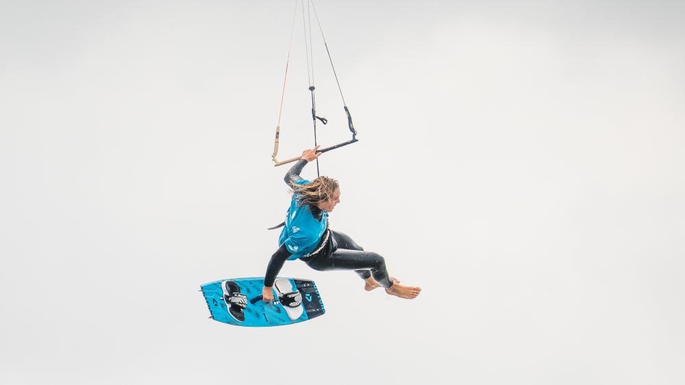 EIn Junge mit blonden langen Haaren in Neopren hängt an Seilen in der Luft. Ein Seiner Hand ein Board.